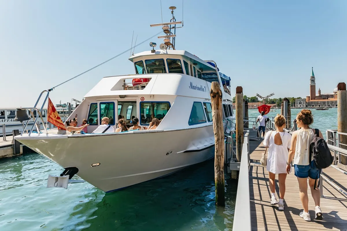 Panoramic tour boat moored near Torcello's grassy foreshore with the brick basilica behind, part of a Murano Burano Torcello boat tour from Venice, Italy